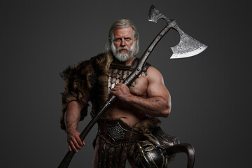 Aged bearded Viking warrior dressed in fur and light armor, holding a two-handed axe, against a...
