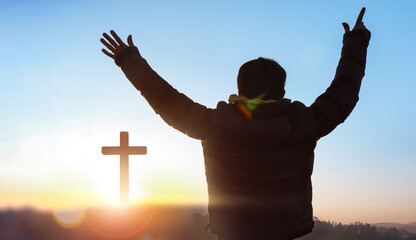 Silhouette of a man praying before a cross at sunrise background.