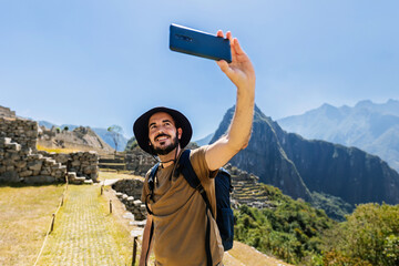 Young adult man taking selfie with phone camera at Machu Picchu. Joyful male tourist enjoying vacation in Peru, South America. Travel and vacation concept.