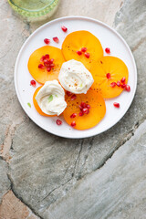 White plate with sliced persimmon, mozzarella cheese and pomegranate, above view on a grey granite background, vertical shot with space