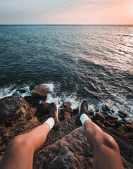 POV view - male tourist (man) sitting on the edge of cliff above the ocean. Man with legs above the sea watching amazing sunset on the horizont.