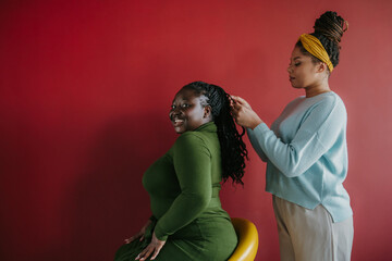 Confident African hairdresser braiding hair to a beautiful young smiling woman on red background