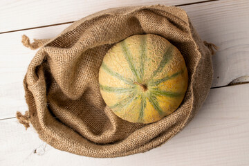 One fragrant organic melon in a jute bag, close-up, on a white wooden table, top view.