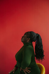 Side view of beautiful curvy African woman with braided hair sitting on the chair on red background