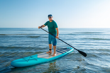 A middle-aged man in shorts and a T-shirt stands on a SUP board with a paddle near the sea.