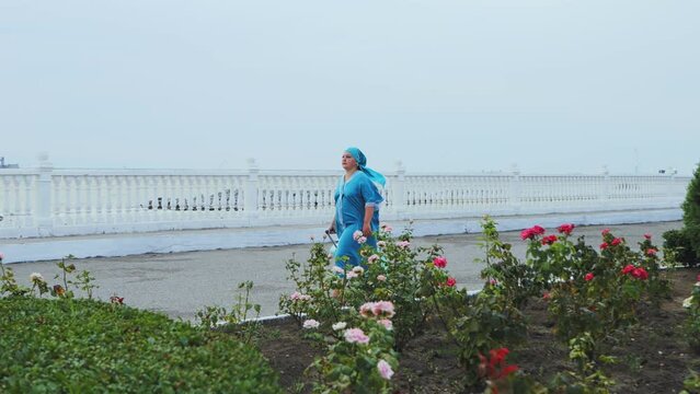 A woman in a turquoise dress and headscarf walks early in the morning with a suitcase along the embankment against the backdrop of the sea past a green alley. Overall plan