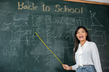 Back to school concept. Asian female teacher smiling with wooden stick pointing to blackboard at school in classroom, Happy beautiful young woman standing hold pointer to back board