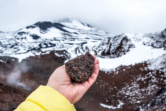 Tourist hand holding lava stone on Mount Etna crater, Sicily, Italy. Warm volcanic lava rock formation. Landscape of craters slopes covered with snow, clouds and smoke