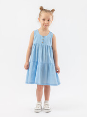 A fashionable 5-year-old girl in a summer blue sundress and white sneakers with a hairstyle stands and looks expressively at a white background. Hands are lowered. Photo.