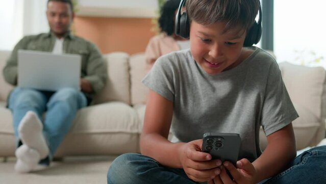 Little boy son playing video game on mobile phone in headphones on floor happy multiracial family using diverse devices at home mom dad working online on laptops background child kid play smartphone