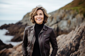 Portrait of a smiling woman in her 50s sporting a stylish leather blazer against a dramatic coastal...
