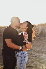 Happy smiling  Couple drinking vine at Sand Dunes near the Beach.  Young happy Bearded muscular  man  in White shirt kissing and hugging beautiful woman at sunset on a beach