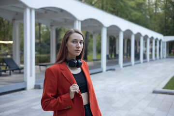 Modern young girl student posing in red jacket with headphones and looking at the camera against the backdrop of white columns receding into the distance in city park. Tourism, relaxing, lifestyle