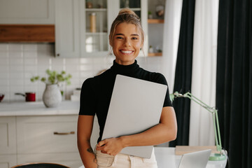 Beautiful young woman holding laptop and smiling at camera while standing at home office
