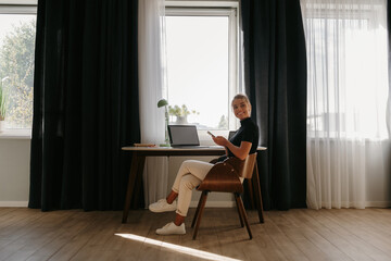 Fototapeta premium Happy young businesswoman working at home while sitting at the desk in front of the window