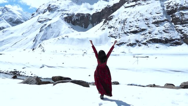 Slow motion shot of teenager Indian girl raising hands in front of snow covered mountains in Lahaul, India. Girl in mountains during frosty Sunny day.