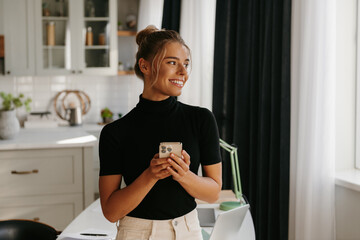 Attractive young woman holding smart phone and smiling while standing at home office