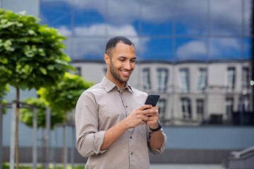 A young successful male businessman in a shirt walks outside an office building, holds a phone in his hands, uses an application on a smartphone, reads online pages, smiles happily