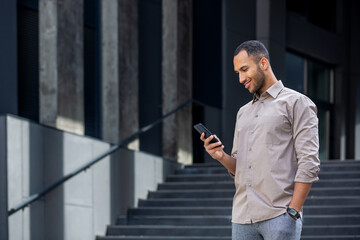 A young successful male businessman in a shirt walks outside an office building, holds a phone in...