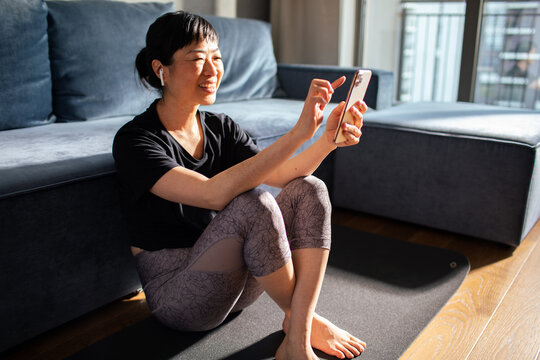 Smiling woman sitting on yoga mat checking smartphone at home