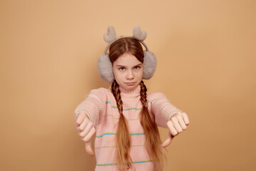 Little upset girl with fluffy headphones showing thumb down on beige background.