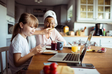 Little girl using laptop during breakfast in the kitchen with mother