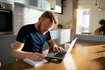 Bearded man using laptop taking notes at home