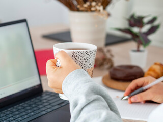 Woman holding a cup of coffee while planning the day in a notebook and looking at laptop. Remote...