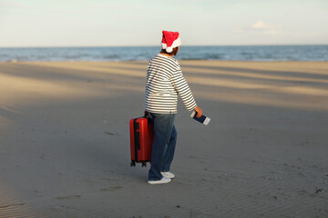 Back view of adult woman in Christmas Santa Claus hat holds red suitcase, passport and boarding pass ticket walks on beach on seashore. Happy New Year 2024 celebration holiday concept.Winter vacation.