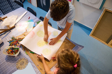 Overhead view of kids holding pencils and drawing on white paper sheet in creative art workshop....