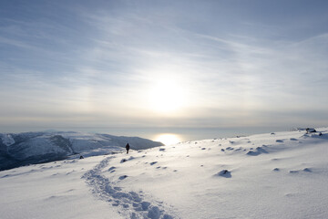 A tourist walks along a snowy mountain slope. Footprints in the snow. There is a solar halo in the sky above the sea. Traveling and hiking in Siberia and the Russian Far East. Magadan region, Russia.
