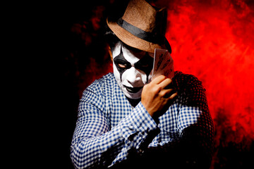 a man's face painted with white and black like joker holding card and smoking cigarettes 