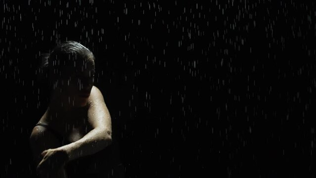 Silhouette slim woman dancing posing under rain at night. Seductive female gymnast dancer wearing black swimsuit, raindrops splashes, water drops roll down sexy body in shower, flashing red light