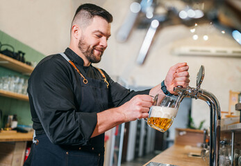 Smiling stylish bearded barman dressed black uniform with an apron tapping fresh lager beer into...