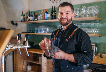 Portrait of happy smiling bearded barman dressed in a black uniform with an apron wiping the beer...
