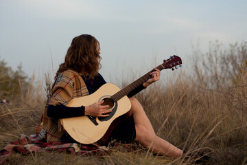 beautiful young smiling woman playing melody on acoustic guitar while sitting in field among tall...