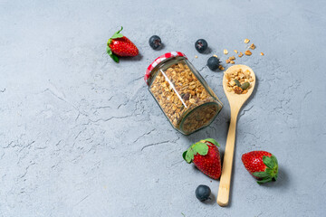 Granola in a glass jar on a concrete table with a wooden spoon, strawberries and blueberries....