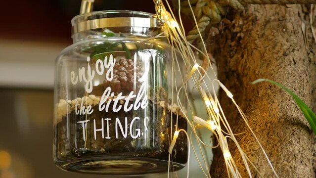 Glass jar with text quote Enjoy the Little Things hanging on a white rope on a tree branch. LED light garland. Halloween decoration to attract guests to restaurant. Rovinj, Croatia - November 14, 2023