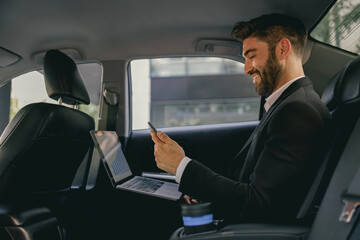 Handsome businessman in suit using personal computer and using mobile phone in taxi - obrazy, fototapety, plakaty