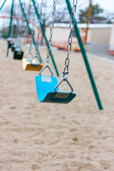 A row of multicolored swings on a playground, with a large sand-covered play area, and portable buildings in the background.