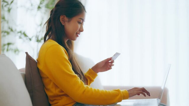 Young asian woman sitting on sofa in living room, makes online banking payments through the internet from bank card on smartphone. Shopping online on mobile phone with credit card