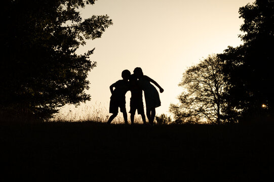 Black shadow silhouette of three kids standing together in the forest, holding each other on a sunset sky and trees background. Friendship or happy summer adventure concept photo.