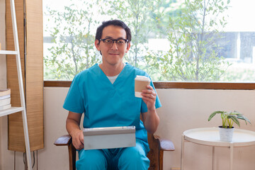 Smiling nurse using digital tablet in employee lounge.  Happy nurse resting on couch in lounge area and drinking cup of coffee.