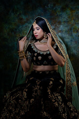 beautiful indian woman in brown bridal lehenga with green dupatta over head and heavy jewellery and bangles sitting on a chair in luxury background holding  veena ( harp)