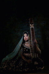 Obraz premium beautiful indian woman in brown bridal lehenga with green dupatta over head and heavy jewellery and bangles sitting on a chair in luxury background holding veena ( harp)