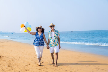 Senior indian couple with colorful balloon in hands walking together at the beach. Enjoying vacation, holiday at beach. Copy space. - obrazy, fototapety, plakaty