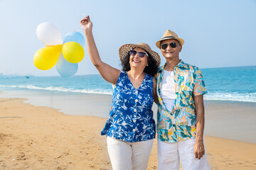 Senior indian couple with colorful balloon in hands walking together at the beach. Enjoying vacation, holiday at beach. Copy space. Retirement life. - obrazy, fototapety, plakaty
