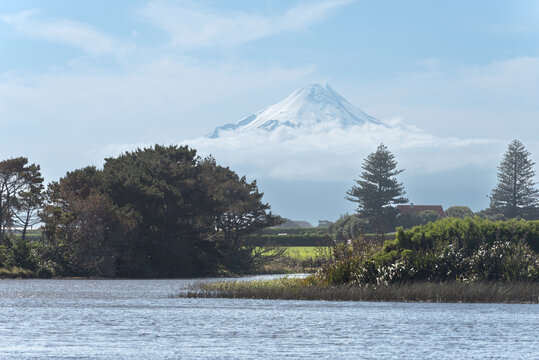 Distant view of snow-capped Mount Taranaki peeking through low clouds with a rural landscape and Lake Lowell's in the foreground.