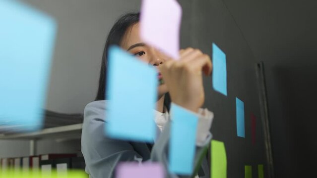 Creative young Asian woman thinking and planning a startup business with paper stickers on a transparent board.