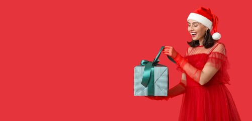 Young woman in Santa hat opening Christmas gift on red background with space for text